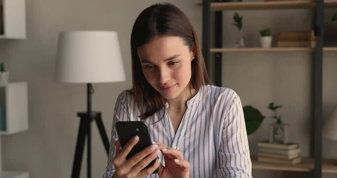Happy attractive millennial woman using mobile phone, swiping photos in dating application, communicating distantly in social networks, checking email, reading news, playing games on cellphone indoors