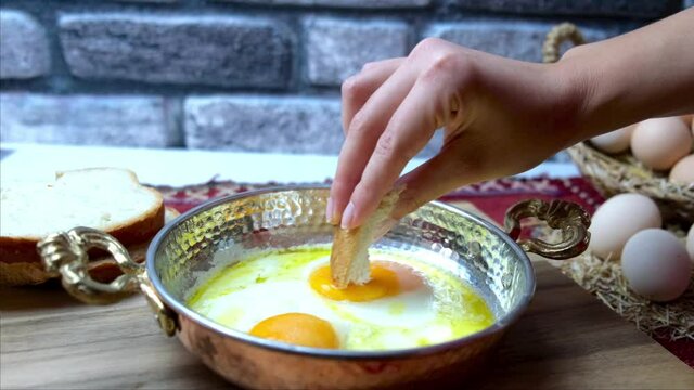 Fried eggs in a copper pan for breakfast, a woman dips bread into an egg.