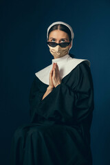 Medieval young woman as a nun in vintage clothing and golden face mask praying on dark blue background. Concept of comparison of eras, fashion, ad, healthcare. Royal person protected from covid.