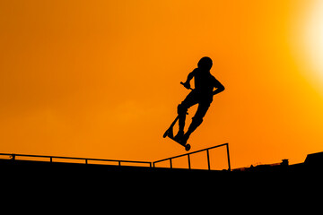 Unrecognizable teenage boy silhouette showing high jump tricks on scooter against orange sunset sky at skatepark. Sport, extreme, youth, urban culture, freestyle, outdoor activity concept