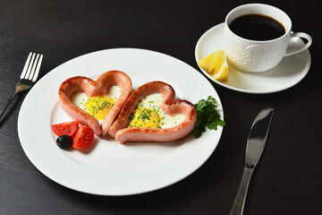 Scrambled eggs in the form of heart on a white plate with sausages, tomatoes, greens and coffee with lemon.