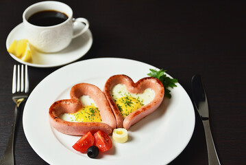 Scrambled eggs in the form of heart on a white plate with sausages, tomatoes, greens and coffee with lemon.