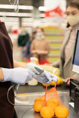 Gloved hands of young female cashier in uniform scanning pack of fresh oranges