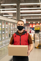 Young courier in uniform and protective mask holding box with fresh products