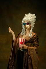 Portrait of medieval young woman in vintage clothes, golden face mask with 3D-eyewear, popcorn on dark background. Royal person protected from covid. Concept of comparison of eras, modern, fashion.