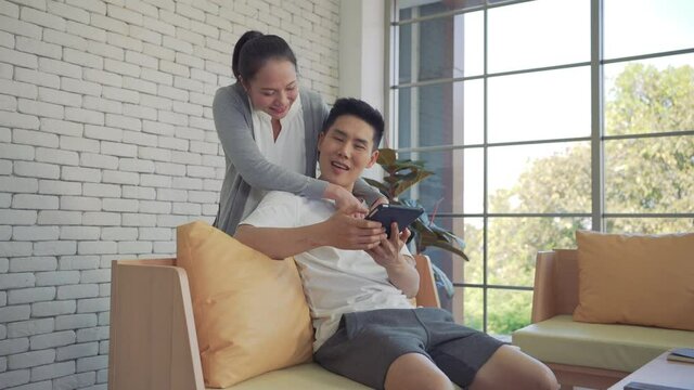 Young asian couple holding tablet together, hugging each other, sitting on a sofa in the luxury contemporary living room next the window at home. Filling their home with love and laughter.