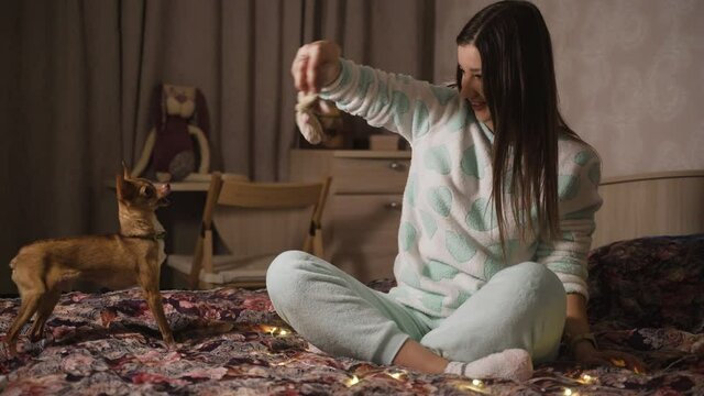 Young girl with long hair plays with dog at home. Brunette and her pet puppy are sitting on the bed in evening