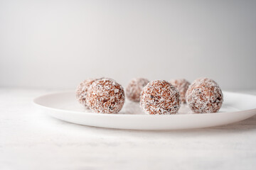 A white plate with round sweets made of dates and coconut shavings on a light wooden background