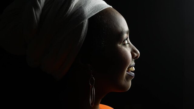 low key portrait of african german woman wearing ethnic white turban and an orange dress turning to camera smiling in front of black background and strong side light giving strong contrast