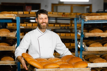 A baker holds a tray with fresh hot bread in his hands against the background of shelves with fresh bread in a bakery. Industrial bread production