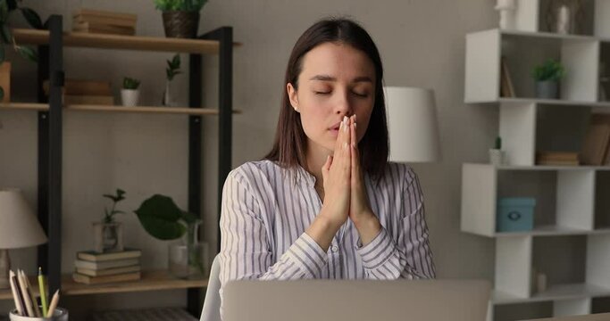 Focused young pretty woman saying worship with folded hands, waiting for professional test results, feeling hopeful at workplace. Mindful religious female worker mediating with closed eyes in office.