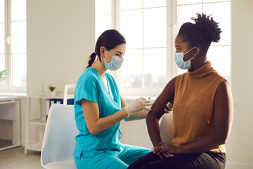 Young African American woman sitting at doctor's office and getting a flu shot in her arm. Nurse in medical face mask holding syringe and giving female patient modern Covid 19 vaccine injection - obrazy, fototapety, plakaty