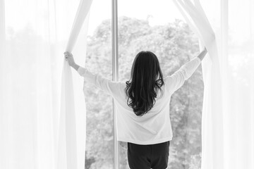 Black and white of Young woman stand open white curtains at the window in morning after waking up in bedroom. Happy female opening window curtains at home. Woman standing by bedroom window.
