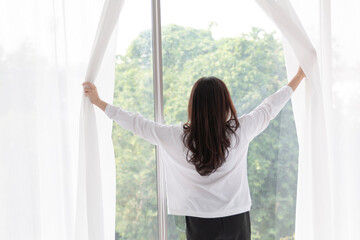 Young woman stand open white curtains at the window in morning after waking up in bedroom. Happy female opening window curtains at home. Woman standing by bedroom window opening curtains.