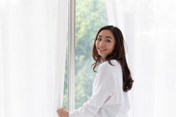 Young woman stand open white curtains at the window in morning after waking up in bedroom. Happy female opening window curtains at home. Woman standing by bedroom window opening curtains.
