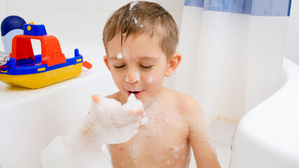 Cheerful laughing boy having bath and blowing soap foam from hand in camera. Concept of child hygiene and health care at home. Kids having fun and playing