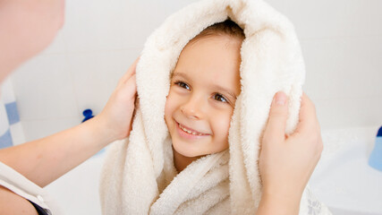Portrait of cute smiling boy with wet hair wiping and drying with soft towel after having bath and looking on caring mother. Concept of child hygiene and health care at home.