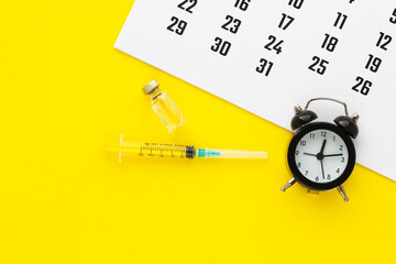 Glass medicine vial with liquid and syringe, calendar and alarm clock on yellow background. Medical...