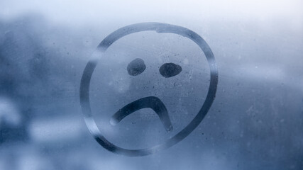 Hand drawn sad emoji on foggy glass window background. Unhappy face sign close-up. Selective focus...