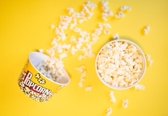 Two full popcorn buckets, one tipped over and surrounded by many popcorn on a yellow background
