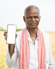 Smiling old Indian farmer standing in front of farmland by holding mobile while looking into to...