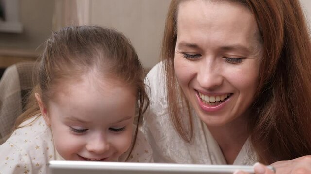 A mother with a small child communicates via the tablet display remotely and laughs. Happy mother and daughter play together with a gadget while lying in bed. Family life. Self-isolation at home