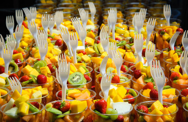 Fresh fruit salad (kiwi, mango, strawberry etc.) arranged in plastic cups with forks on a market stall. Selected focus. Healthy fast food concept.