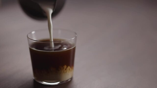 Slow motion milk pour into tumbler glass with coffee on walnut table