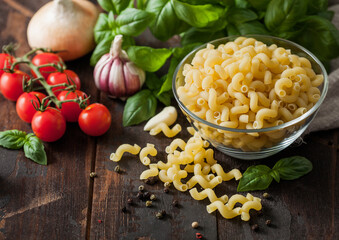 Raw maccheroni elbows pasta in glass bowl with oil and garlic, basil plant and tomatoes with pepper on wooden background.