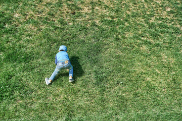 Fototapeta na wymiar Playful little boy in blue cap goes up steep hill slope covered with lush green grass in spring park on sunny day backside view