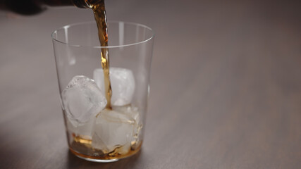 pour cola over ice cubes into tumbler glass on walnut table with copy space