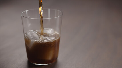 pour cola over ice cubes into tumbler glass on walnut table with copy space