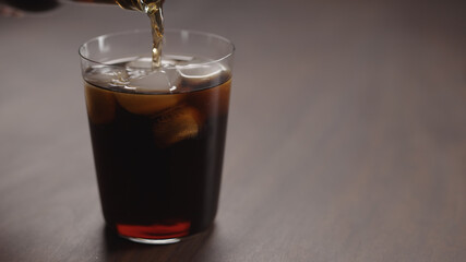 pour cola over ice cubes into tumbler glass on walnut table with copy space