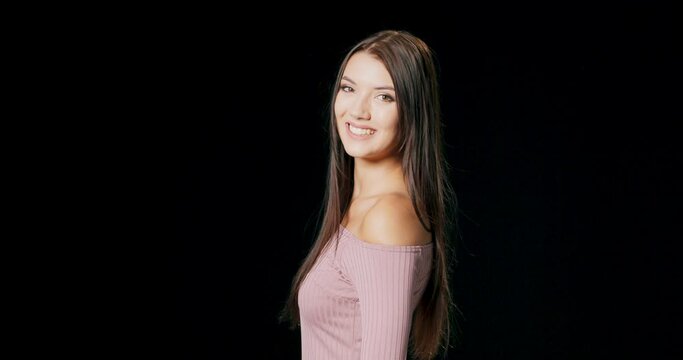 Portrait of a young beautiful woman wearing pink dress turning around, smiles and looking at you. She has dark long hair and wonderful big green eyes. Black background.