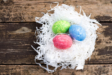 Three easter colors eggs are arranged on nest of white paper straw. Easter is main event for believers, timed coincide with resurrection of Jesus Christ. Nest on wooden background. 