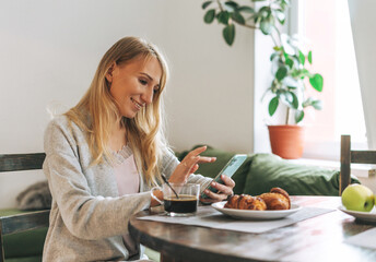 Beautiful blonde young woman having breakfast and using mobile in living room at home