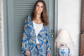 attractive young smiling brunette woman with long hair in silk cardigan with blue indian paisley pattern stays against grey door inside