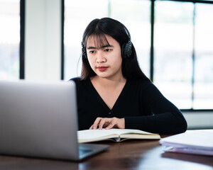 Smiling Asian girl student wears wireless headphones write on the notebook to study language online watch and listen to the lecturer, webinar via video call e-learning at home, distance education