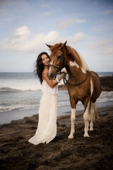 Obraz premium Woman leading horse by its reins. Horse riding on the beach. Love to animals. Asian woman wearing long white dress. Travel concept. Copy space. Bali