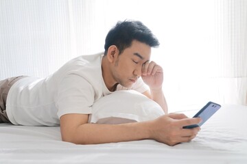 Sleepy Young Asian man looking smart phone in bed