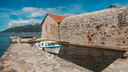 The man is clinging to a stone wall of a medieval church in Montenegro. A small private boat at the pier and beautiful mountains in the background. Island Gospa od Milosti in Montenegro
