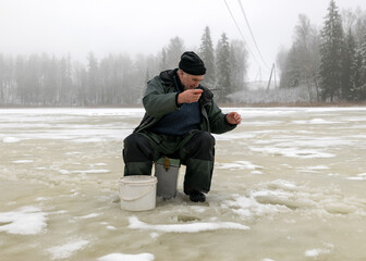 landscape with a man in winter who sits on the ice next to a hole and holds a small fishing rod. A fisherman is sitting on a frozen lake and fishing, winter