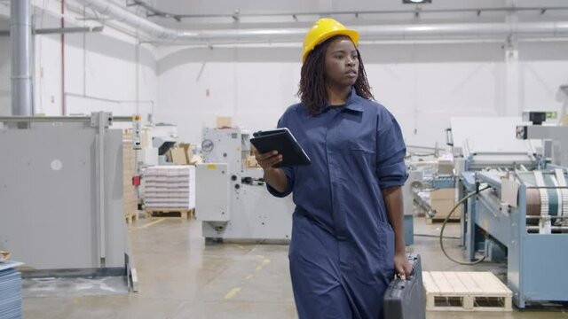 Black female factory engineer with tablet and toolkit box walking on plant floor, monitoring machines work. Dolly shot. Industry and production concept