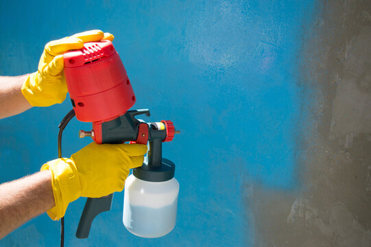 A worker's hands in yellow gloves holds a hand-held spray gun with an overhead compressor. Painting the wall of the house in blue. Household renovation. Copy space. Selective focus