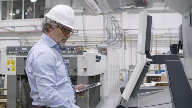 Senior engineer testing industrial machine on plant floor, pressing buttons at control board, monitoring process, using tablet. Production process or machinery concept