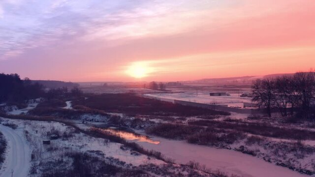 Bright sun in the clear sunset evening sky in frosty weather Bright sun on the horizon and glare on the river thaw. A drone flight over a snow-covered rural landscape to the setting sun on the horizon