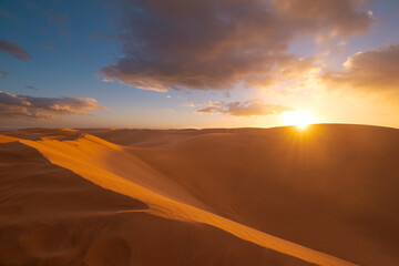 Fototapeta na wymiar Golden sand dunes in desert in Maspalomas. Sunset in the desert, sun and sunrays, beautiful dramatic clouds and blue sky. Gran Canaria, Canary islands, Spain