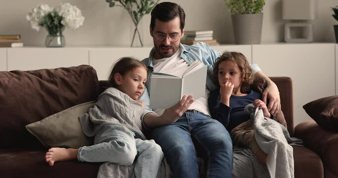 Loving young father reading book interesting fairy tale to little preschool son and daughter, family enjoy leisure time at home relaxing on comfy couch covered with plaid. Children development concept