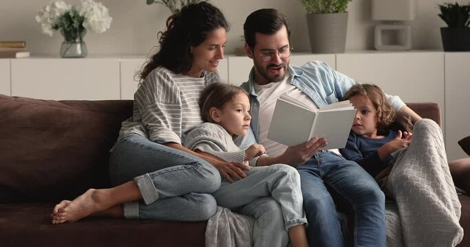 Full family gathered in cozy living room, couple and kids resting on comfy couch reading fairy tale to little curious attentive son and daughter. Development education of children, fun, hobby concept