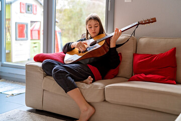 Teenager girl playing guitar sitting on sofa at home in living room. Child hobby and leisure...